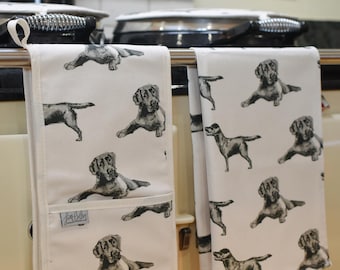 May include: A set of two white kitchen tea towels featuring a black and white illustration of a Labrador Retriever dog. The towels are hanging on a rack in a kitchen.