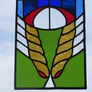 May include: Stained glass panel with a black border, featuring a design of abstract shapes and colors. The panel includes blue, red, white, green, and yellow-gold hues. The design has a central white oval shape, red arch, and two yellow-gold, textured, wing-like shapes.