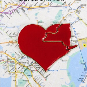 May include: Red heart-shaped pin with a gold outline of the state of New York. The pin is on a map of New York City.