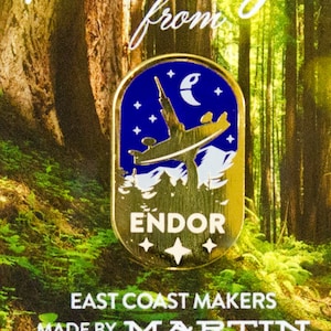 Pode incluir: Pin de esmalte dourado com uma nave espacial voando sobre uma cena de floresta com uma lua crescente e estrelas. O texto "ENDOR" está impresso em branco no pin. O pin é rodeado por um fundo de floresta verde.