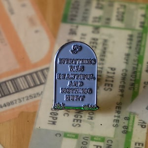 May include: A silver and green enamel pin shaped like a tombstone with the text "Everything was beautiful, and nothing hurt".