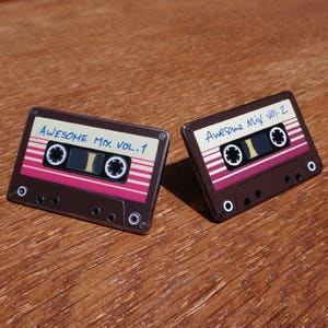 May include: Two brown cassette tape pins with a pink and white stripe design. The text on the tapes reads "Awesome Mix Vol. 1" and "Awesome Mix Vol. 2".