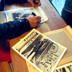 May include: A person signing black and white photographs on a wooden table. One photo features a man in a coat with handwritten text. Other photos are visible, along with a pen and a black marker.