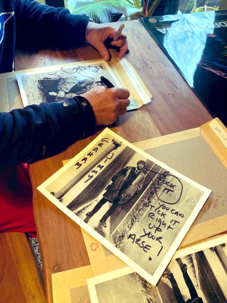 May include: A person signing black and white photographs on a wooden table. One photo features a man in a coat with handwritten text. Other photos are visible, along with a pen and a black marker.