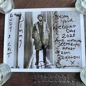 Könnte beinhalten: Schwarz-Weiß-Fotografie eines Mannes in Anzug und Mantel, der in einer engen Gasse steht. Handschriftlicher Text enthält "Enjoy Your Wedding Day 2023" und Unterschriften. Das Foto ist von kleinen Glaskerzenhaltern umgeben.