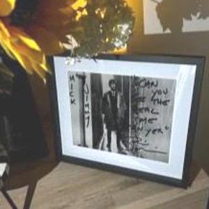 May include: Framed black and white photograph featuring a man standing in front of a wall with graffiti. The text "Can you feel the real me yet?" is visible on the wall. The frame is black with a white mat.