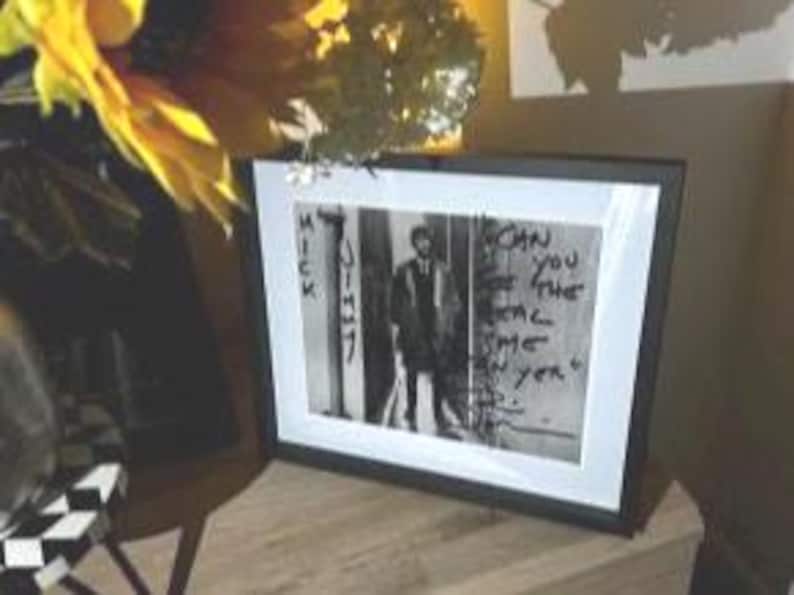 May include: Framed black and white photograph featuring a man standing in front of a wall with graffiti. The text "Can you feel the real me yet?" is visible on the wall. The frame is black with a white mat.