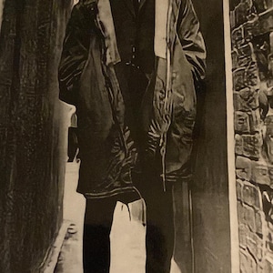 May include: A black and white photograph of a person wearing a dark suit, tie, and overcoat, standing in a narrow alleyway. The person is wearing black shoes. The alley has brick walls on one side.