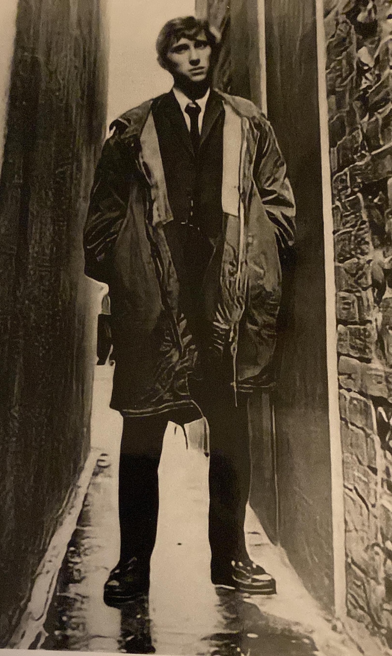May include: A black and white photograph of a person wearing a dark suit, tie, and overcoat, standing in a narrow alleyway. The person is wearing black shoes. The alley has brick walls on one side.