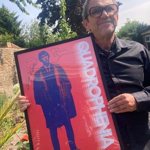 Może przedstawiać: Oprawiony plakat ze słowem QUADROPHENIA w białym, ułożonym tekście na czerwonym tle. Plakat przedstawia niebiesko-fioletową ilustrację osoby w długim płaszczu. Plakat ma podpisy w lewym górnym rogu.
