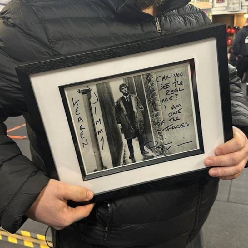 May include: Black and white framed photograph featuring a man in a coat standing in an alley. The frame is black, and the photograph has handwritten text that reads "Can you see the real me? I am one of the faces."