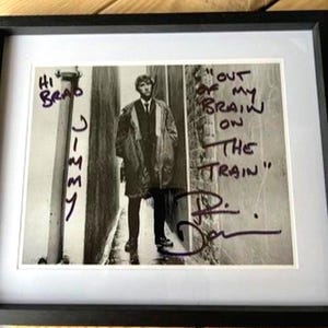 May include: Framed black and white photograph of a man in a coat, standing in an alley. Handwritten text includes "Hi Brad Jimmy" and "Out of my brain on the train". Signed by the man in the photo.