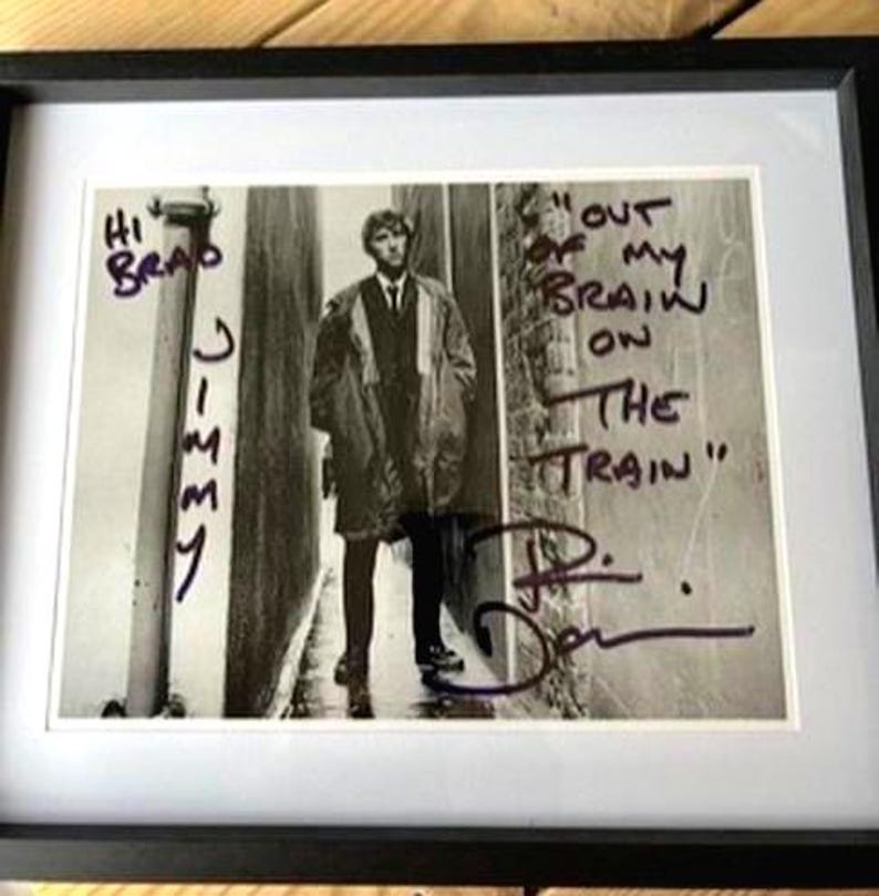 May include: Framed black and white photograph of a man in a coat, standing in an alley. Handwritten text includes "Hi Brad Jimmy" and "Out of my brain on the train". Signed by the man in the photo.