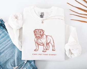 May include: White sweatshirt featuring a brown illustration of a bulldog. The text "CAN I PET THAT DAWG?" is printed below the dog. A silver necklace with a small pendant is visible.