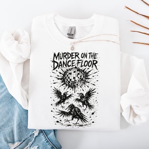 May include: White sweatshirt with the text "MURDER ON THE DANCE FLOOR" above a black graphic of a disco ball and three crows. The design is in black ink. A silver necklace is draped on the sweatshirt.
