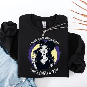 May include: Black sweatshirt with a graphic of a witch holding a potion, with the text "I don't curse like a sailor, I curse like a witch." A silver necklace is draped across the sweatshirt. The sweatshirt is paired with blue jeans.
