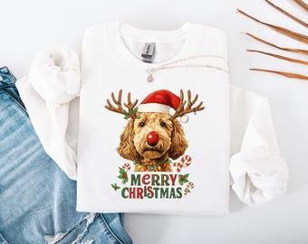 Può includere: Felpa bianca con un design natalizio. Presenta un cane con corna di renna, un naso rosso e un cappello di Babbo Natale. Le parole "MERRY CHRISTMAS" sono stampate in verde e rosso sotto il cane.