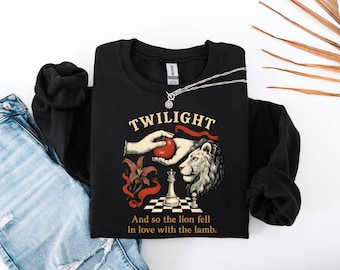 May include: Black sweatshirt with the word "TWILIGHT" above a graphic of a hand holding a red apple, a lion's head, and a chess piece. The text "And so the lion fell in love with the lamb" is below the graphic. A silver necklace is on top of the sweatshirt.