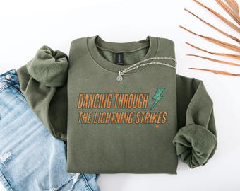 May include: An olive green sweatshirt with the text "DANCING THROUGH THE LIGHTNING STRIKES" in orange and teal. A silver necklace with a pendant is draped across the sweatshirt. The sweatshirt is folded next to a pair of blue jeans.
