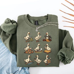 Puede incluir: Sudadera verde oliva con un diseño de dibujos animados de gansos cosiendo. Los gansos se muestran con herramientas de costura y tela, creando un tema lúdico. La sudadera está doblada sobre unos vaqueros. Una cadena de plata está encima.