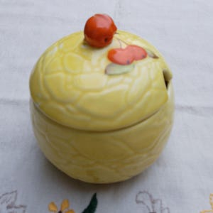 May include: A yellow ceramic sugar bowl with a lid featuring a red cherry design. The bowl has a textured surface and a small spout for pouring.