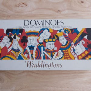 May include: A rectangular box of dominoes with the text "DOMINOES" and "Waddingtons" printed on it. The box features a colorful illustration of playing card figures. The box also includes a book of games, a cribbage board, and pegs.