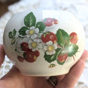 May include: A small, round, off-white ceramic bowl. The bowl is decorated with a strawberry design, featuring red strawberries, green leaves, and white flowers. The bowl is held in a person's hand.