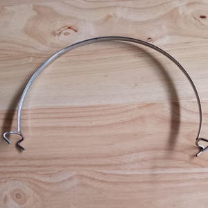 May include: A curved, silver-colored metal band with hooked ends. The band is arched and appears to be made of a single piece of metal. The hooks at each end are small and curved. The item is set against a light brown wooden surface.