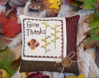 Puede incluir: Un cojín decorativo con las palabras "Give Thanks" bordadas en un cuadrado blanco. El diseño incluye mazorcas de maíz y una cesta de cosecha de otoño. El cojín tiene un botón marrón y está rodeado de hojas de otoño.