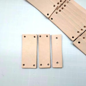Peut inclure: Trois morceaux rectangulaires de cuir brun clair avec deux trous chacun. Le cuir est lisse et a une surface légèrement texturée.