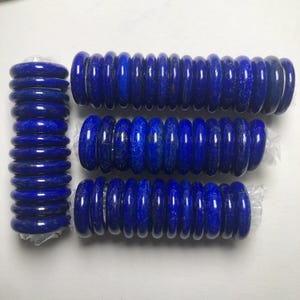 Puede incluir: Una colección de anillos de piedra lapislázuli de color azul intenso dispuestos en cuatro pilas distintas. Los anillos son lisos, pulidos y de forma cilíndrica, con tonos azul oscuro naturales y sutiles motas de pirita dorada en la piedra.