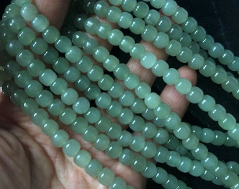 Pode incluir: Um fio de contas de jade verde claro, cada conta é redonda e ligeiramente achatada. As contas são enfiadas em um fio transparente e estão dispostas em um padrão simétrico.