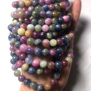 Puede incluir: Una pila de pulseras de piedras preciosas multicolores con cuentas redondas. Las pulseras están hechas de piedras preciosas azules, verdes, rosas y amarillas.