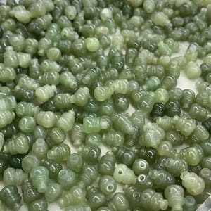 May include: A pile of small, translucent green beads. The beads are cylindrical with an uneven surface. The colour ranges from pale to deep green, with some beads appearing almost black. The beads are scattered, creating a textured appearance.
