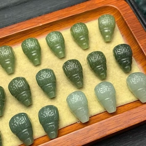 Puede incluir: Una colección de colgantes de jade tallado en varios tonos de verde, dispuestos en una bandeja de madera. Cada colgante tiene forma de calavera estilizada con detalles intrincados. Los colgantes varían en color.