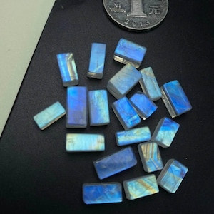 Puede incluir: Una colección de 17 cabujones rectangulares de piedra lunar azul iridiscente. Las piedras son de varios tamaños y tienen una base blanca con destellos iridiscentes azules y dorados.