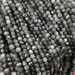 May include: A collection of small, faceted square beads in shades of charcoal grey and silver. Each bead features multiple polished surfaces that reflect light, creating a shimmering, textured appearance across the dense, uniform arrangement.