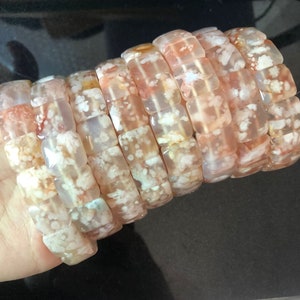 Puede incluir: Una pila de cuentas de ágata de forma cuadrada con un patrón de color blanco y rosa. Las cuentas son translúcidas y tienen un aspecto natural y orgánico.