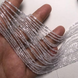 Op de afbeelding: Meerdere strengen kleine, heldere, vierkant geslepen kralen in een trapsgewijze formatie. De kralen zijn transparant en reflecterend, wat een glinsterend effect creëert over de talrijke parallelle lijnen die in een uniform patroon naar beneden vallen.