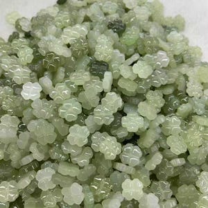 Puede incluir: Un montón de cuentas translúcidas en forma de trébol de color verde claro. Las cuentas tienen un acabado brillante y diferentes tonos de verde. El diseño de trébol de cuatro hojas es claramente visible en cada cuenta, sugiriendo suerte o naturaleza.