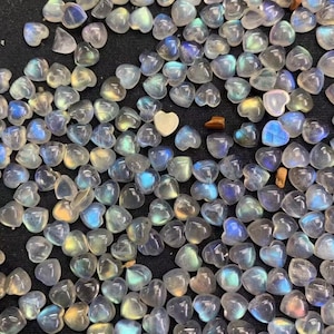 Op de afbeelding: Een verzameling van regenboogkleurige, iriserende maanstenen hartvormige cabochons. De stenen zijn doorschijnend en hebben een melkachtig witte kleur met een regenboogkleurige glans.