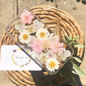 May include: A clear phone case with pressed flowers inside. The flowers are white, pink, and yellow. The text "Blooming in June" is on a white card in the background.