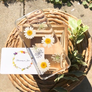 May include: A clear phone case with pressed white daisies and other small white flowers. The case is on a woven basket with a white card that says "Blooming in June".