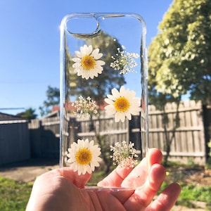 May include: A clear phone case with pressed white daisies and small white flowers. The flowers are pressed inside the case and are visible through the clear plastic.