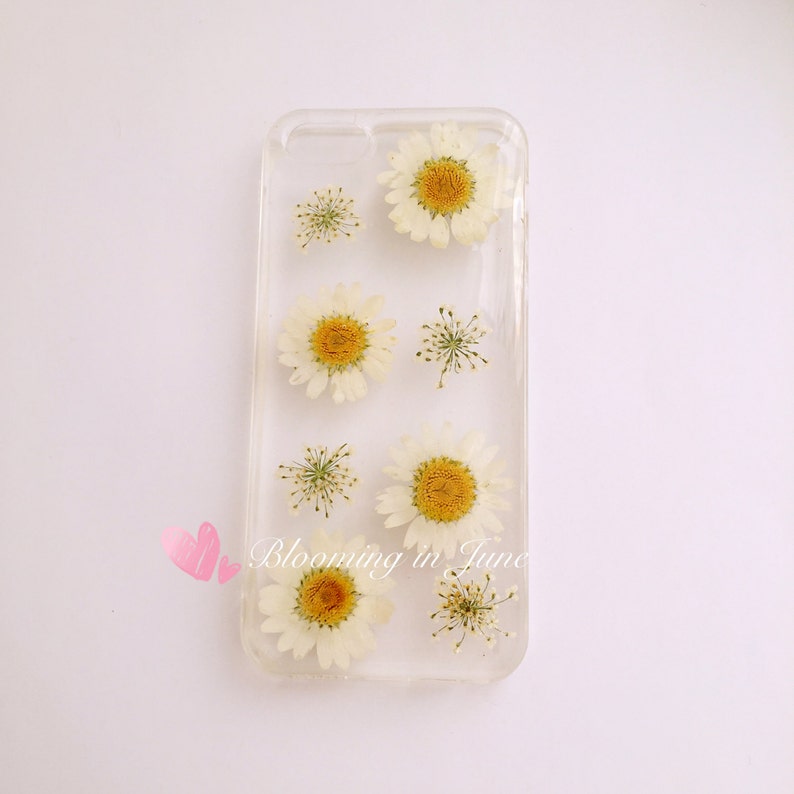 May include: A clear phone case with pressed white daisies and green stems. The text "Blooming in June" is printed on the case.