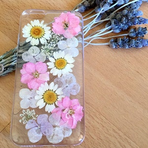 May include: A clear phone case with pressed flowers. The case features white daisies, pink flowers, and light purple flowers.