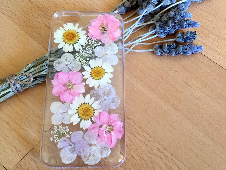 May include: A clear phone case with pressed flowers. The case features white daisies, pink flowers, and light purple flowers.