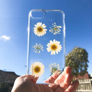 May include: A clear phone case with pressed white daisies and small white flowers. The case is held in a hand against a blue sky background.