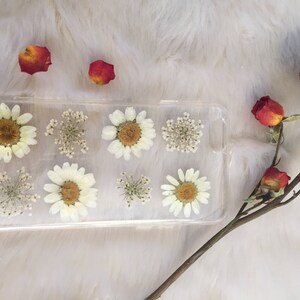 May include: A clear phone case featuring pressed white flowers and greenery. The case is rectangular in shape and decorated with small white flowers and green leaves.
