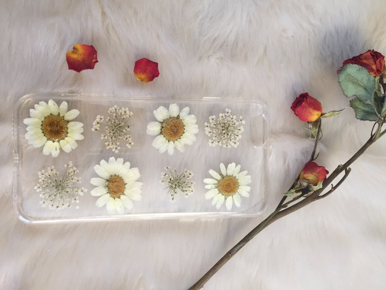 May include: A clear phone case featuring pressed white flowers and greenery. The case is rectangular in shape and decorated with small white flowers and green leaves.